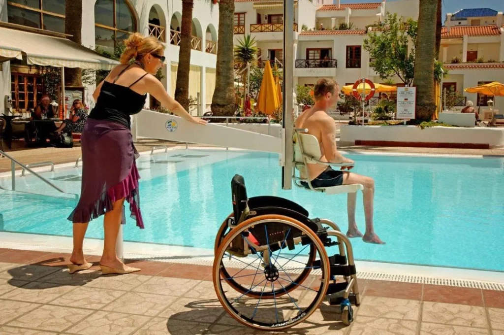 A disabled traveler using a pool hoist lift to enter a swimming pool at Mar y Sol hotel, illustrating essential amenities for accessible winter travel.
